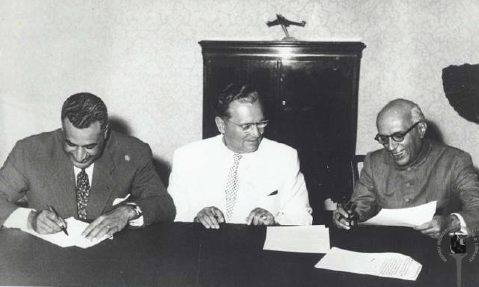 Three men sit at a table, smiling, each looking over a piece of paper. One man is writing something down.
