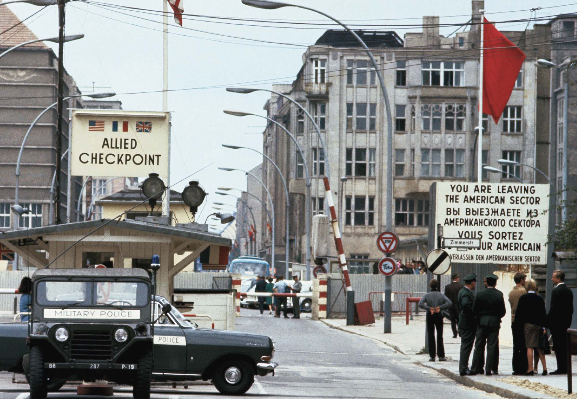 A photograph of a military checkpoint. There is a sign that reads “You are leaving the American sector”, with several people standing in front of it. The checkpoint is a booth, with two military police cars parked in front of it.