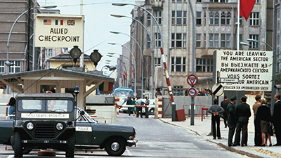 A photograph of a military checkpoint. There is a sign that reads “You are leaving the American sector”, with several people standing in front of it. The checkpoint is a booth, with two military police cars parked in front of it.