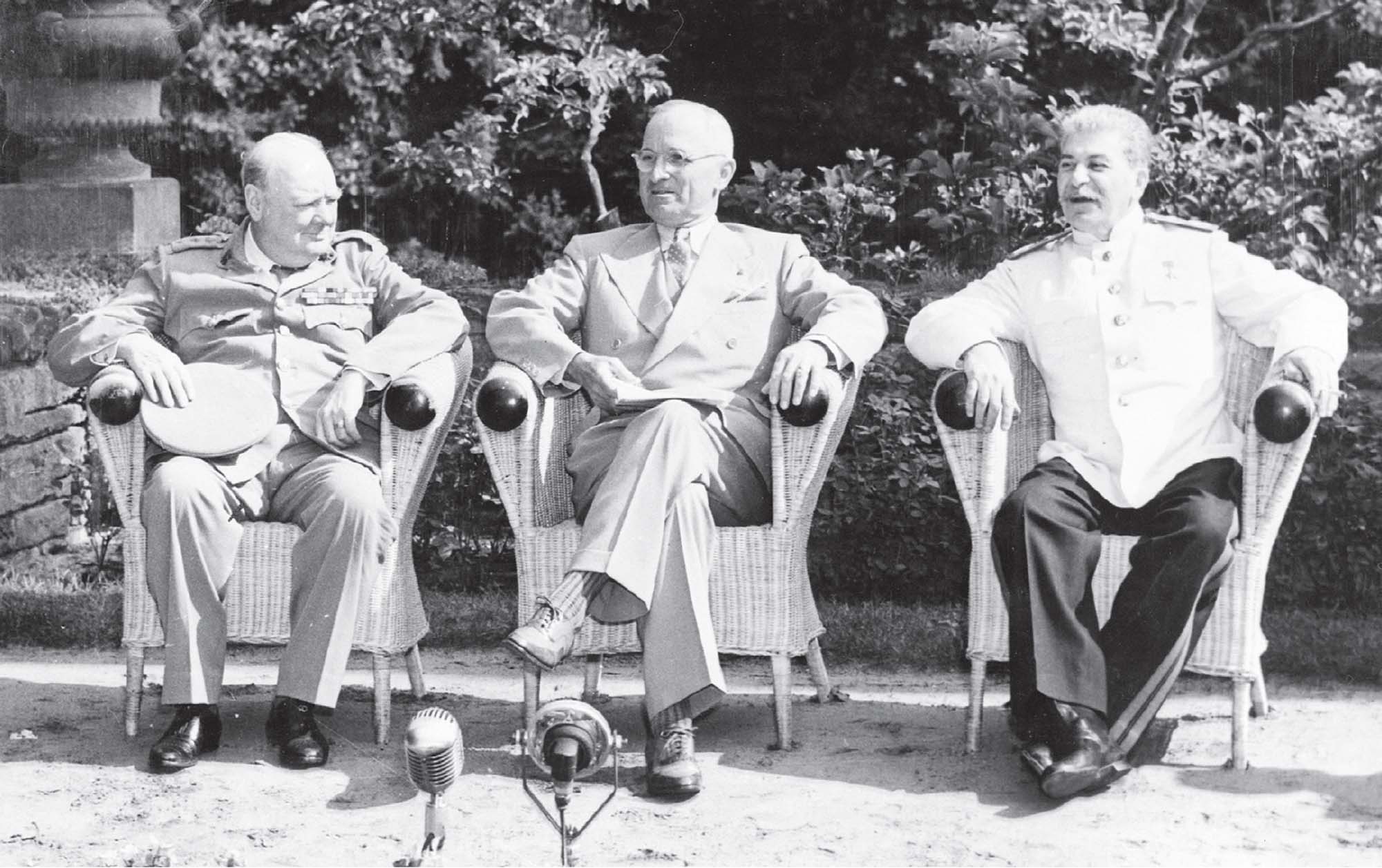 Black and white photo of three older men sitting in wicker chairs outdoors, with microphones in front of them.