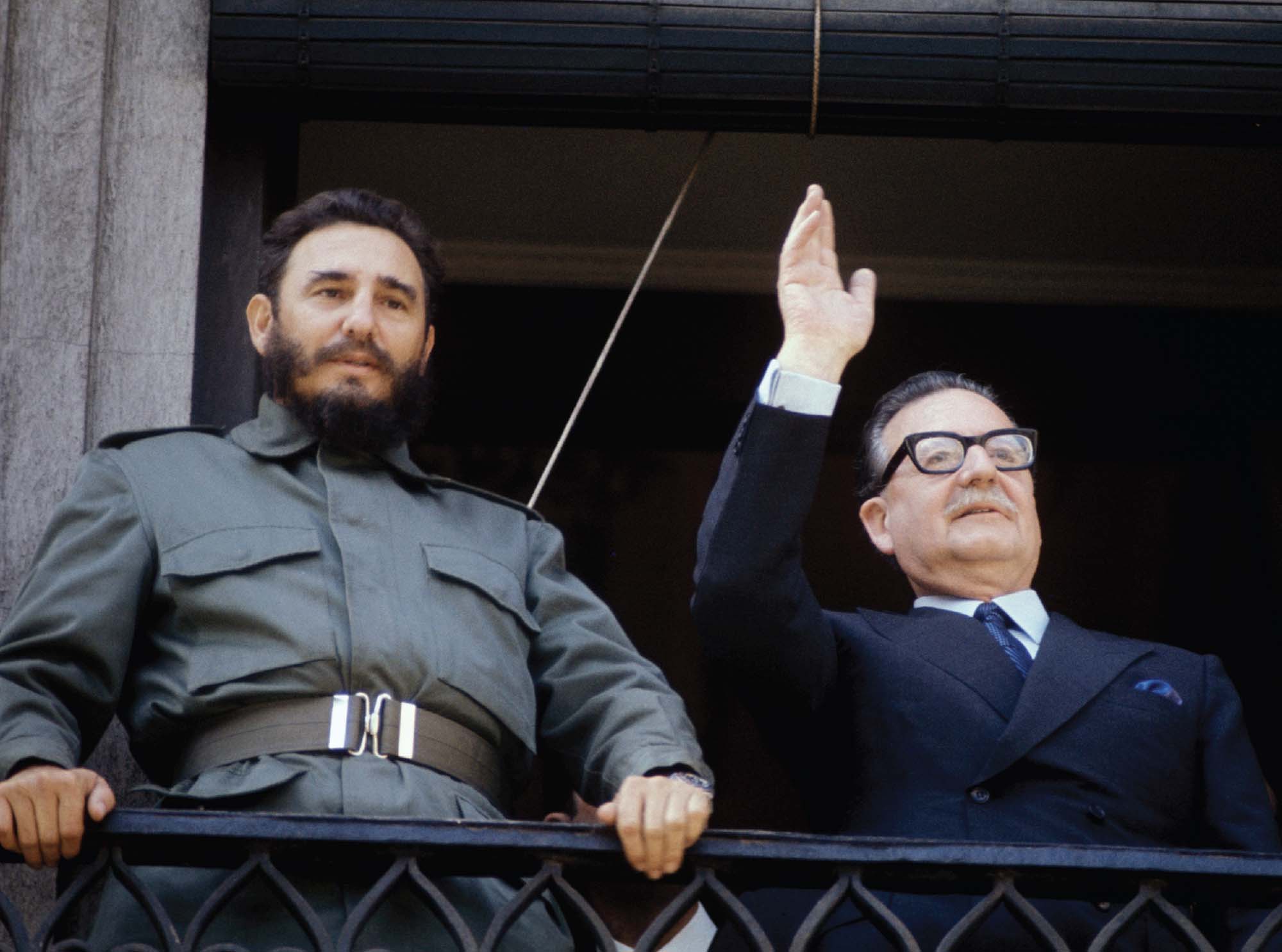 Photo of Cuban premier Fidel Castro in uniform holding onto a railing in front of him and Chilean president Salvador Allende on the right, waving.