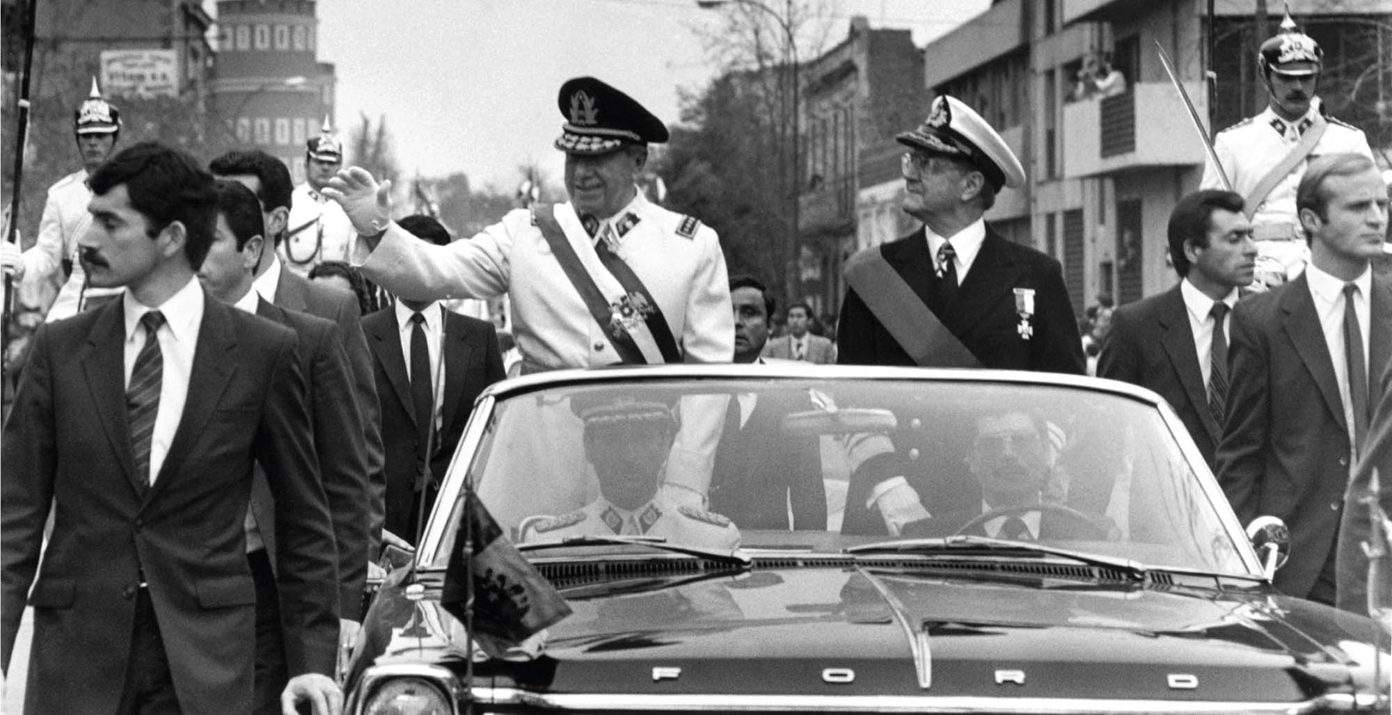 A high-ranking military ruler rides in a convertible waving to supporters, protected by several bodyguards.