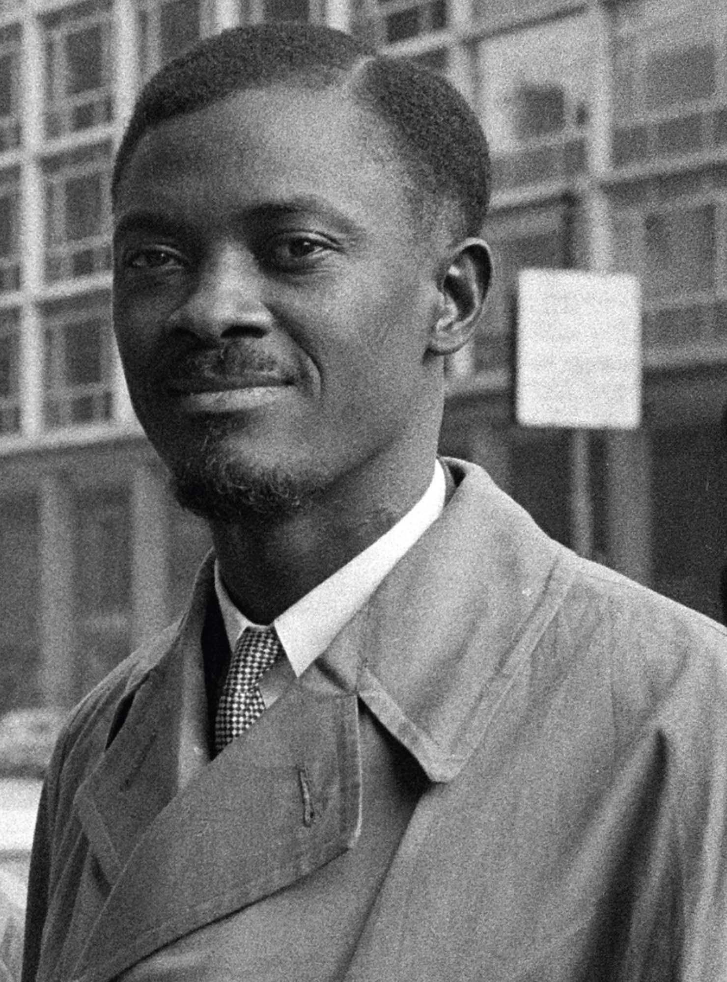 A black and white photo of a man, looking at the camera, standing on the street. He wears a faint smile and is dressed in an overcoat and tie.