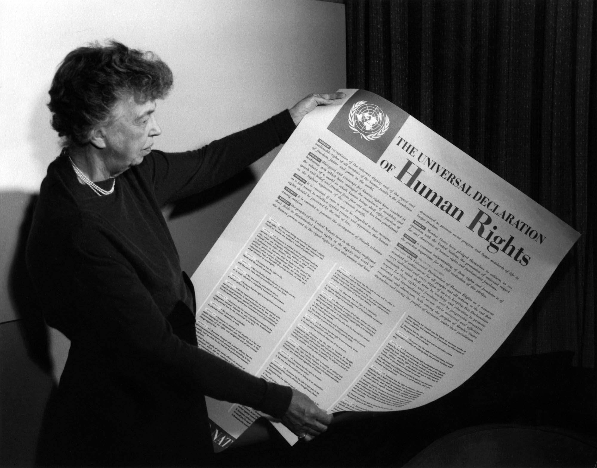 Lady Eleanor Roosevelt holds up a large printed broadsheet of the Declaration of Human Rights.