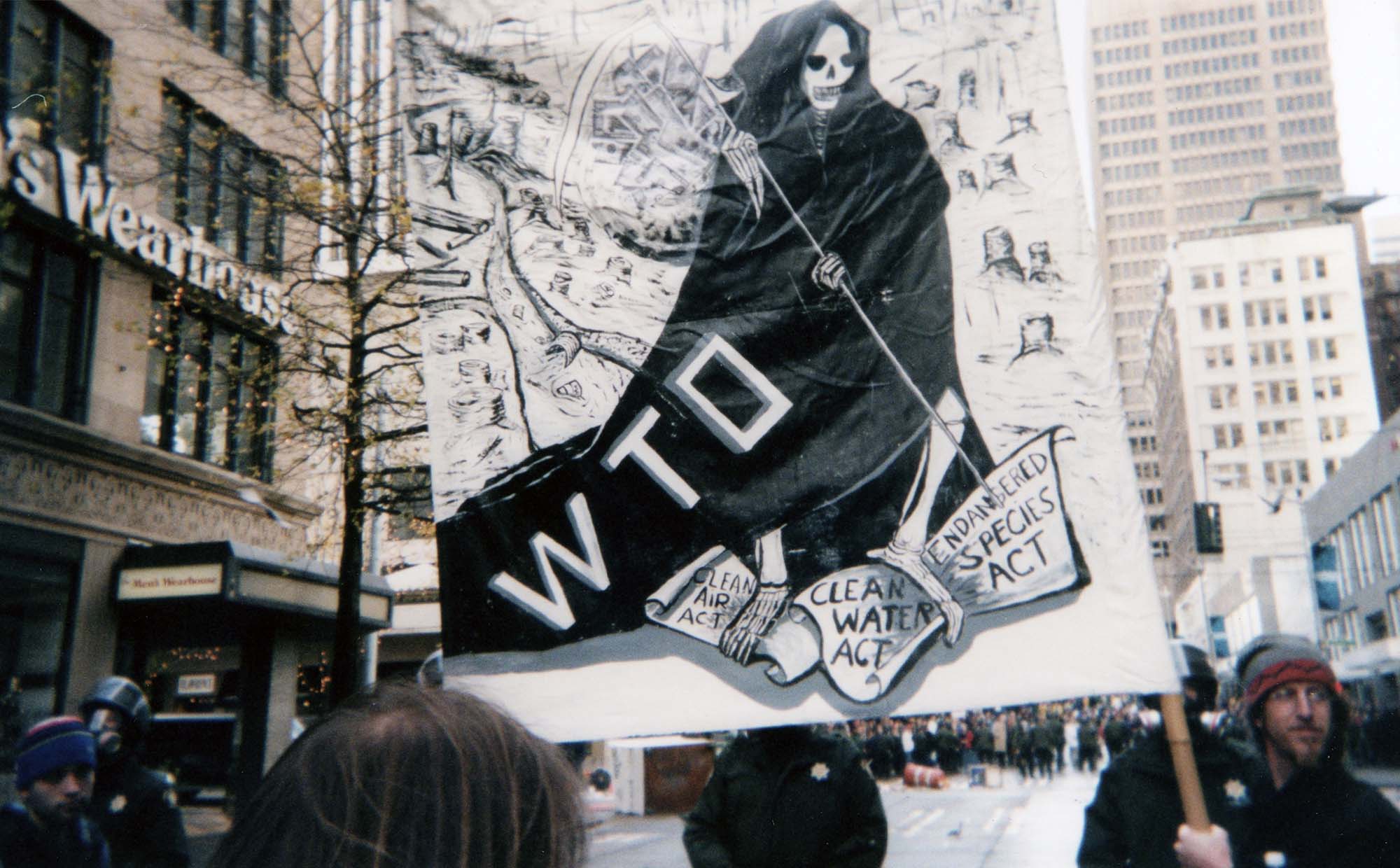 Protestors march and carry banners denouncing the WTO. The banner shows the Grim Reaper standing on regulatory bills for the clean air act, clean water act, and endangered species act .