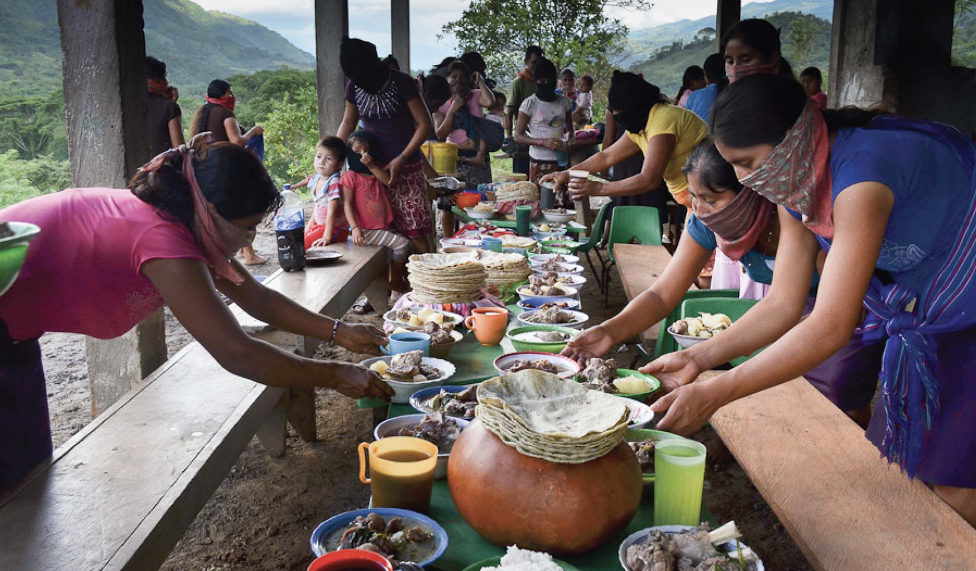 Several people set a table under a wooden shelter. Most are wearing handkerchiefs around their noses and mouths and are filling the table with a large feast. Behind them is a view of a lush, mountainous area.