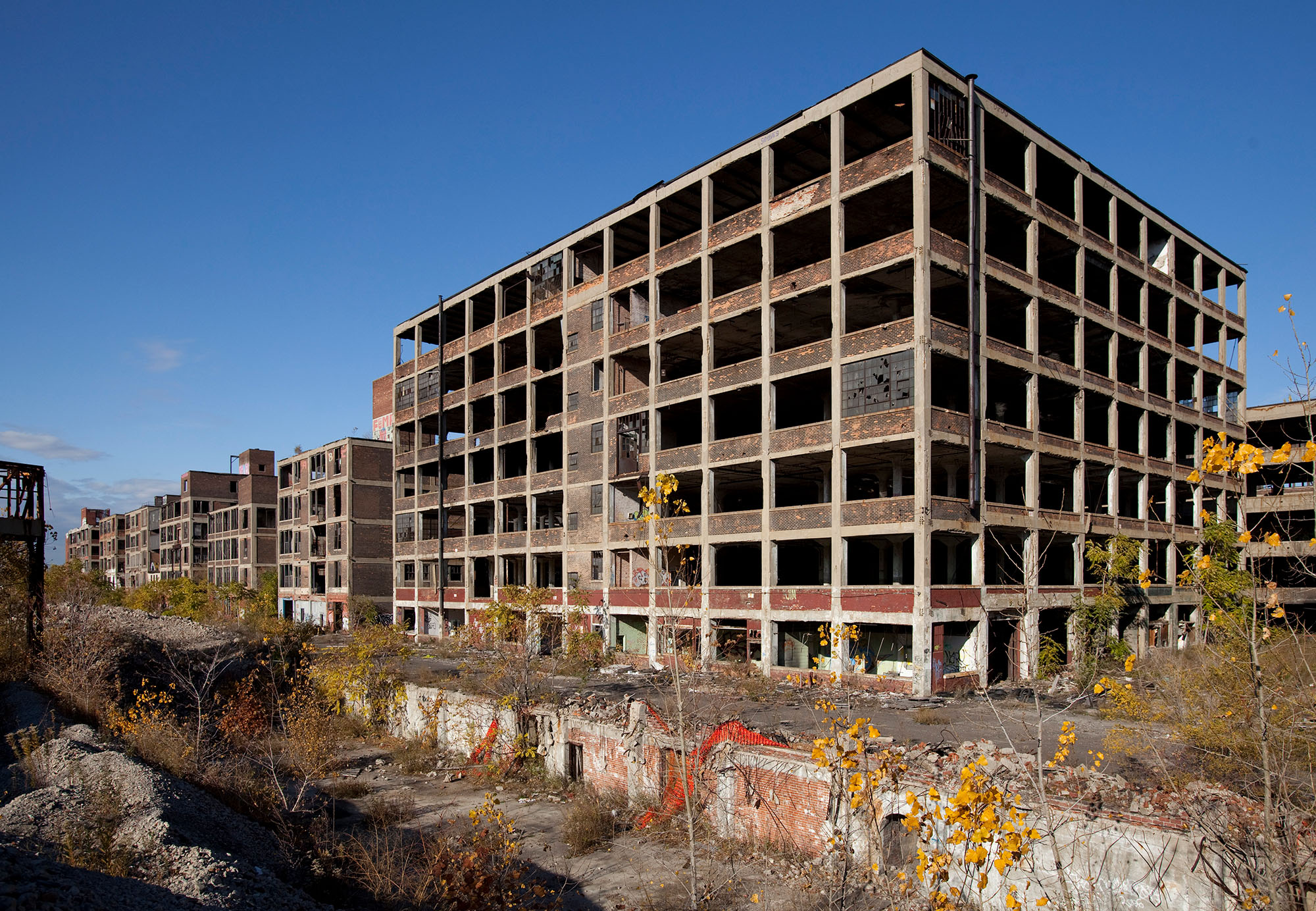 An abandoned automobile factory. The shell of a concrete building with broken windows, overgrown plants, and graffiti. 