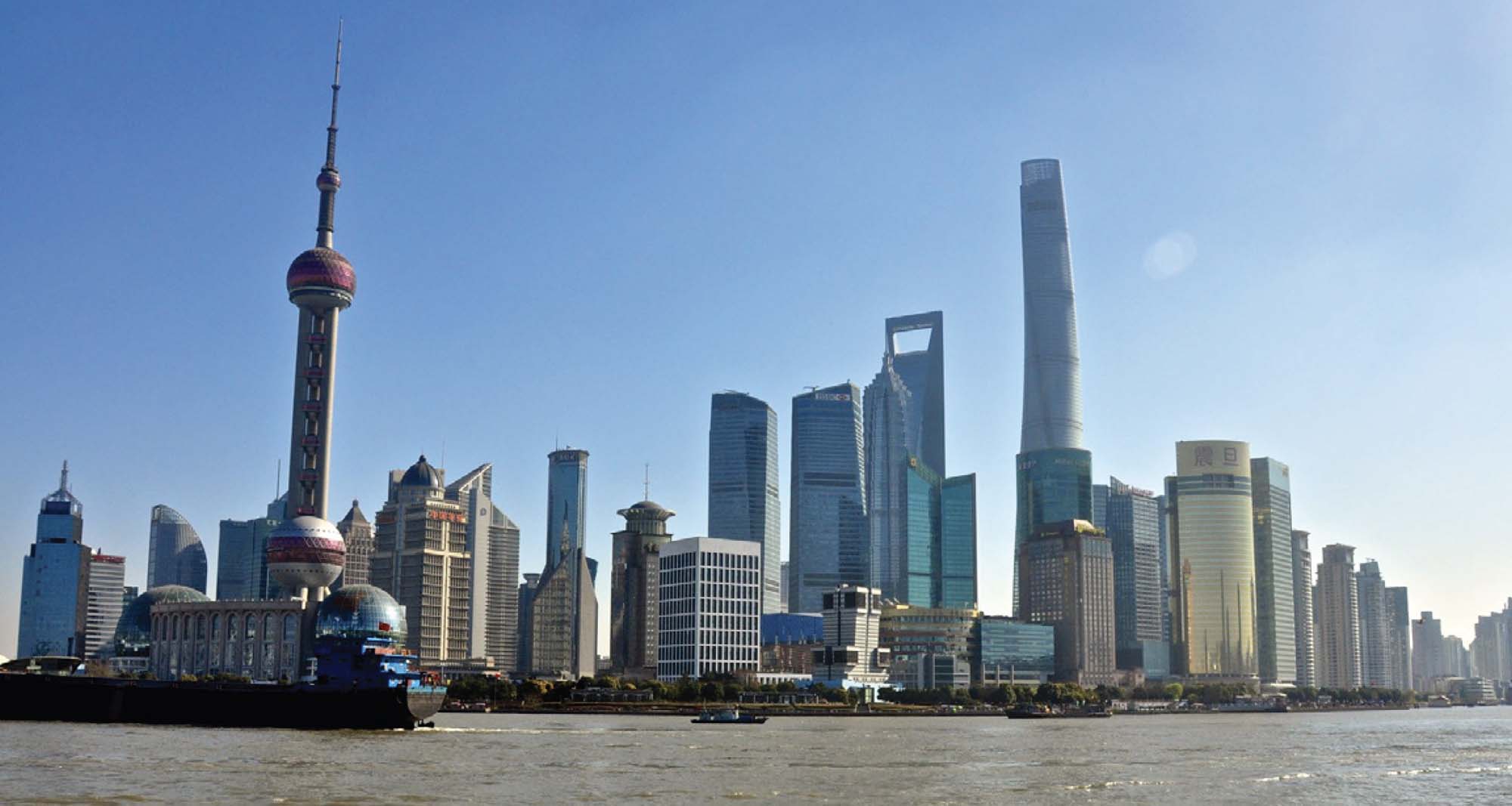 A modern-day photograph shows an impressive skyline, featuring many tall and modern buildings, along the water. A ship is on the water in front of the skyline.