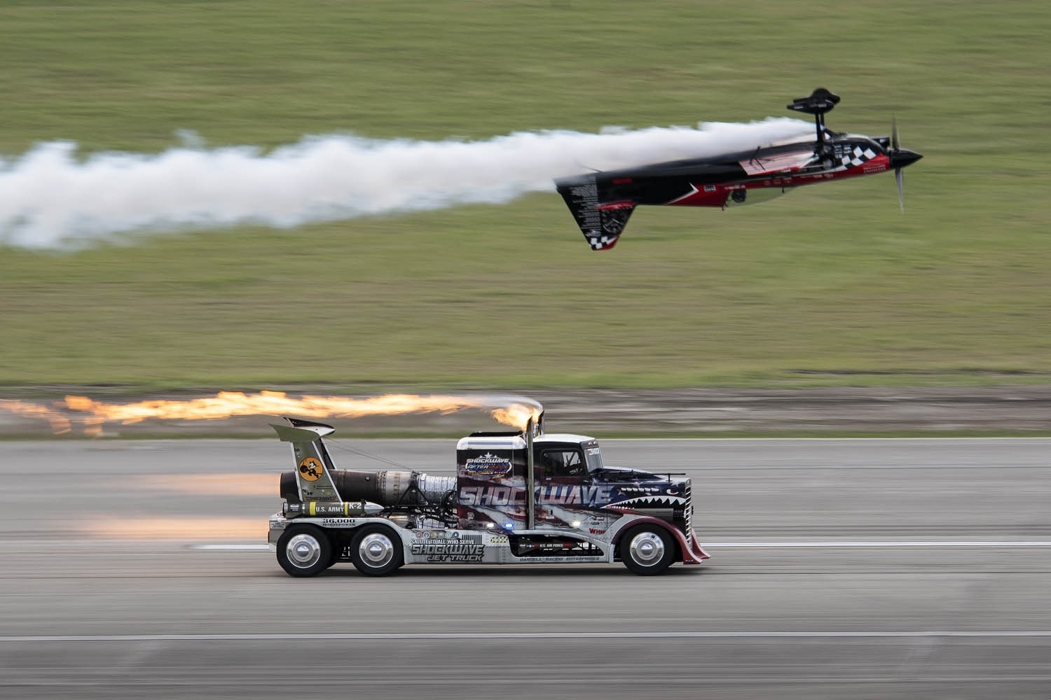 An aerobatic aircraft flies upside down over a fast-moving rocket truck with flames shooting out its engines.