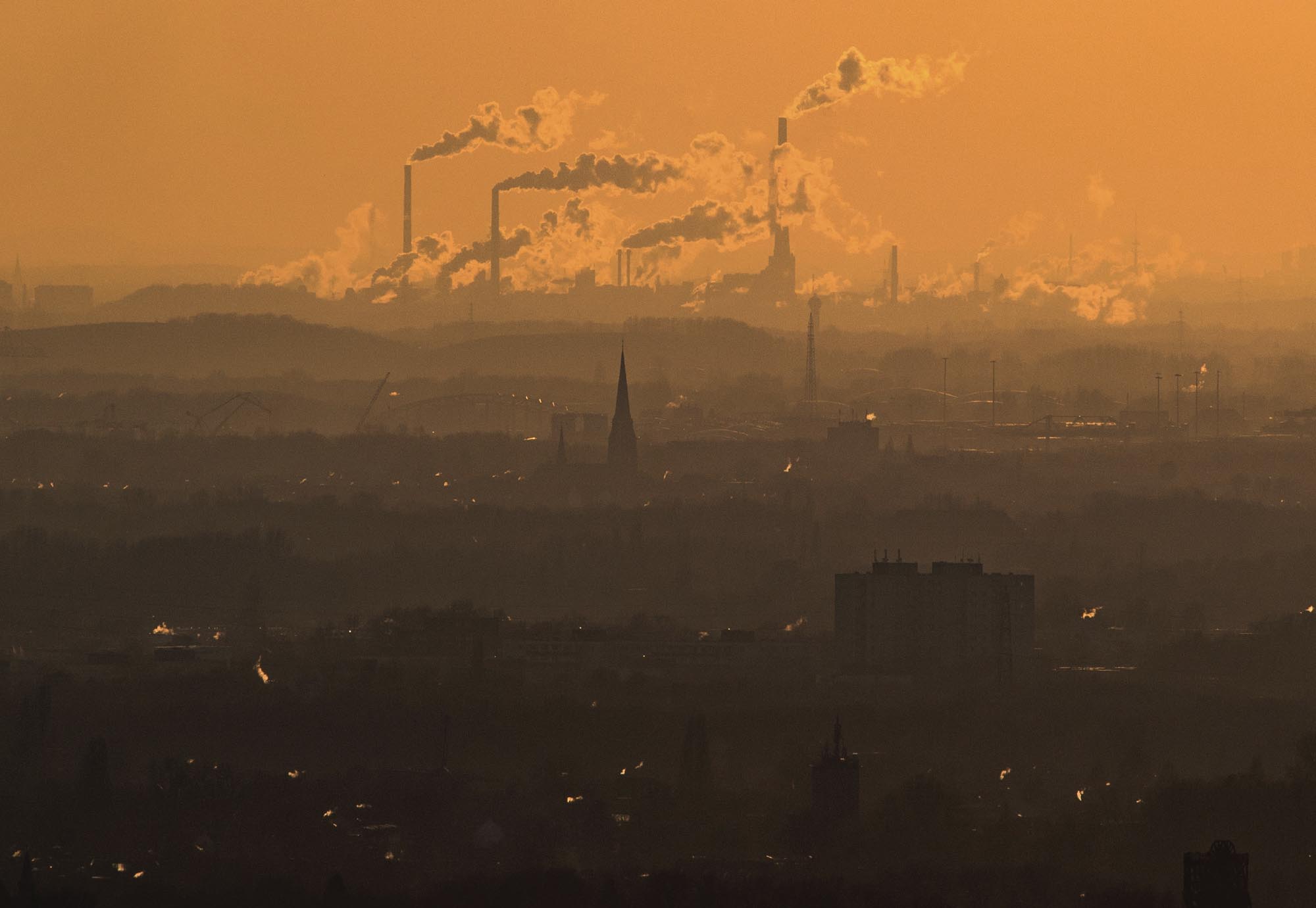 A photograph of a hazy, dark orange sky over an industrial city. Tall smokestacks emit large billows of smoke.