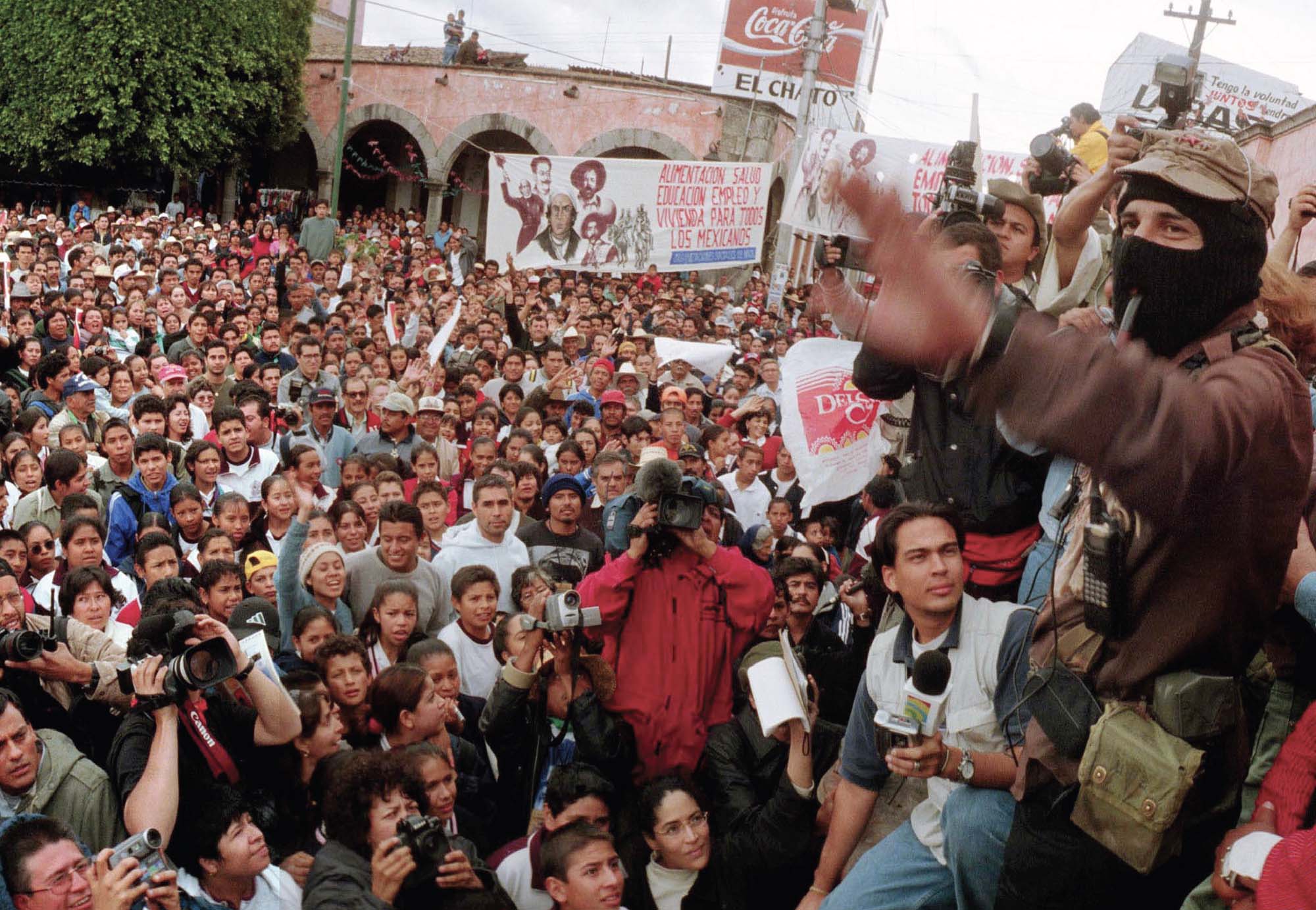 A large crowd gathers at a rally, some people holding up signs and some people holding cameras. Several people stand on a pedestal, one of whom is wearing a face covering, and around them are reporters with microphones.
