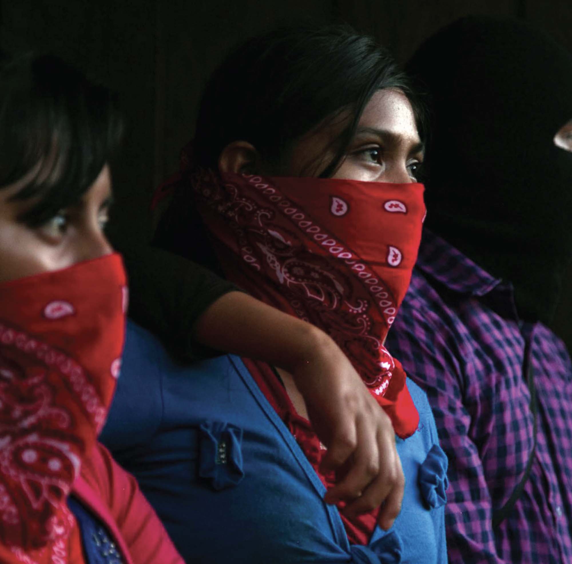 Three people stand in a line. Two people are wearing red bandanas over their noses and mouths, and one person, mostly obscured in the photo has their arm around one of the people wearing red bandanas.