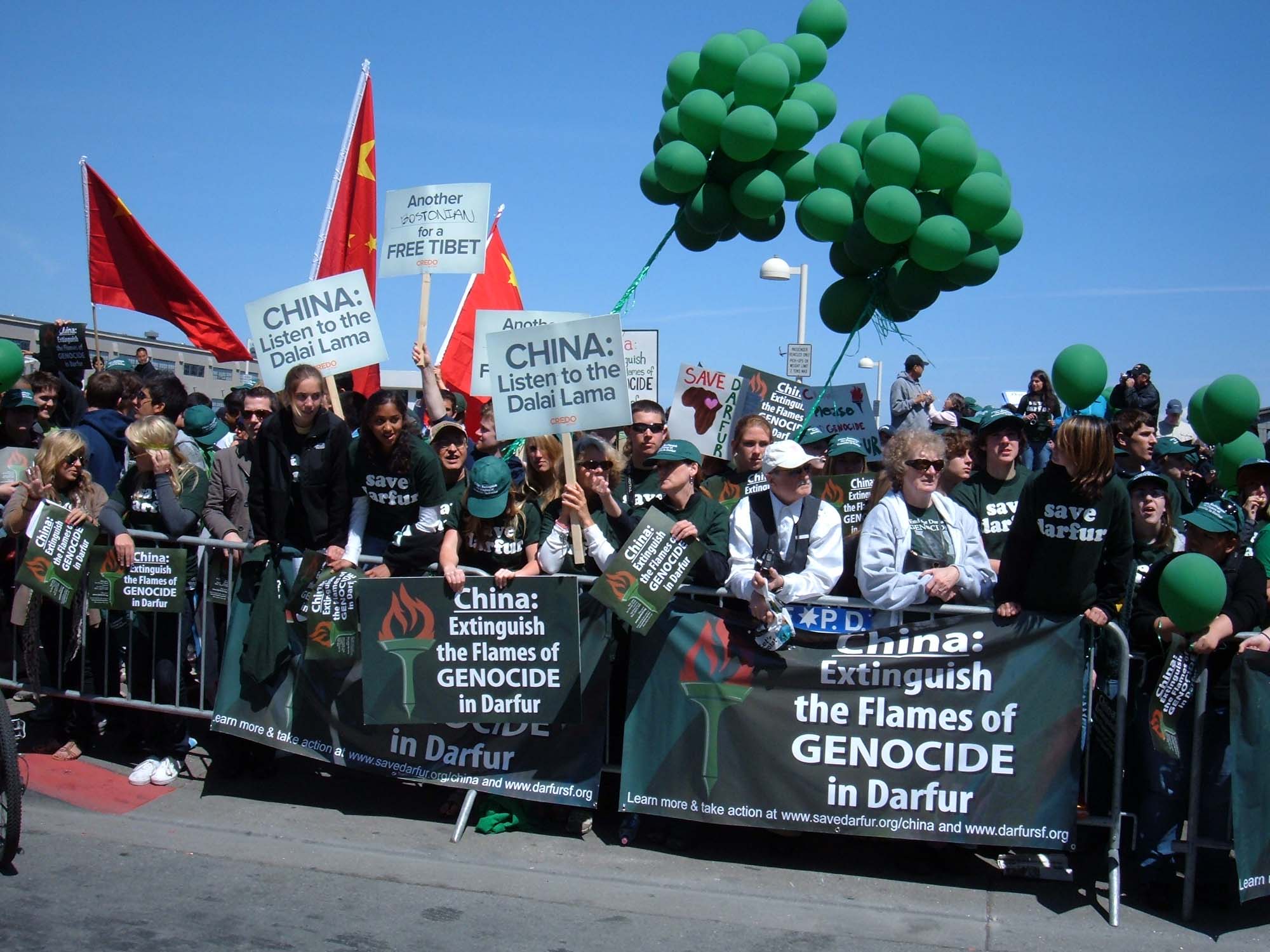 Photograph shows protestors gathered behind metal gates, with signs attached that read “China: Extinguish the Flames of GENOCIDE in Darfur”. People are holding green balloons and signs that read “China: Listen to the Dalai Lama”.