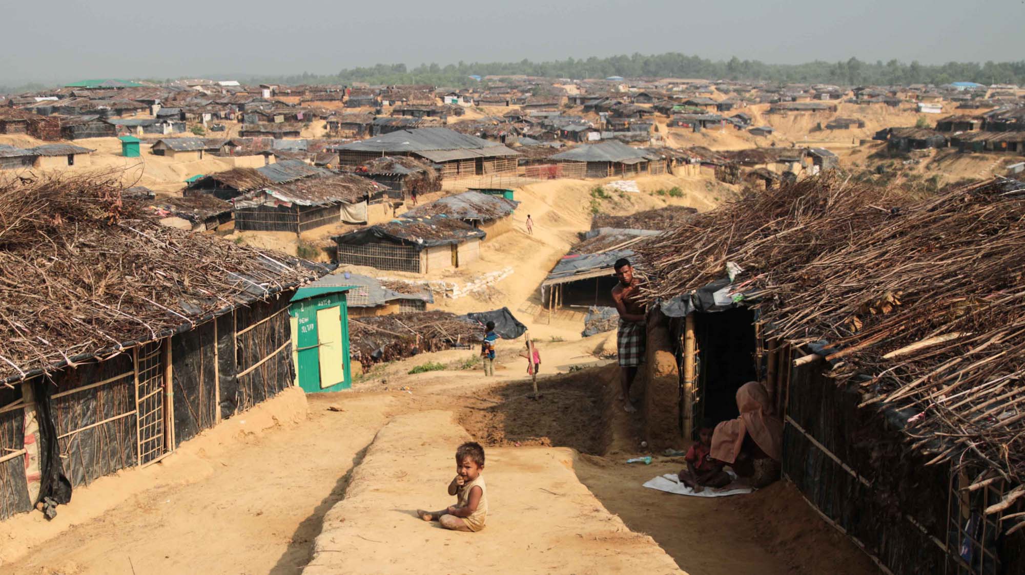 A photograph of a baby, seated on the ground on a narrow walking path between rows of thatched huts.