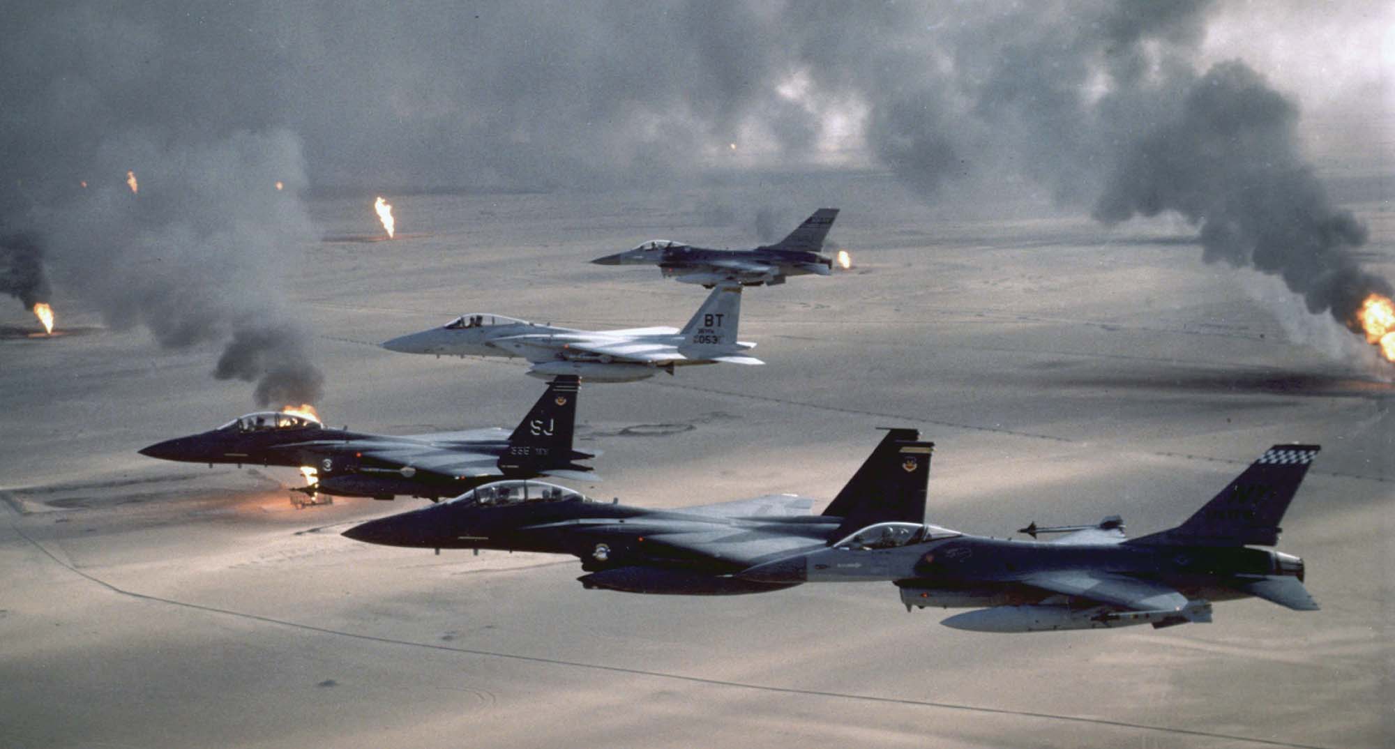 Photo of American military planes flying in a V formation over Iraq’s burning oilfields. Flames and smoke can be seen billowing up into the air from the ground.