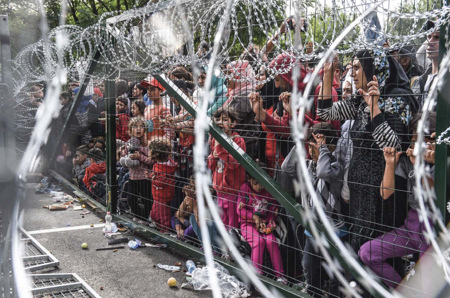 A crowd of refugees being held back behind a metal fence and razor wire.