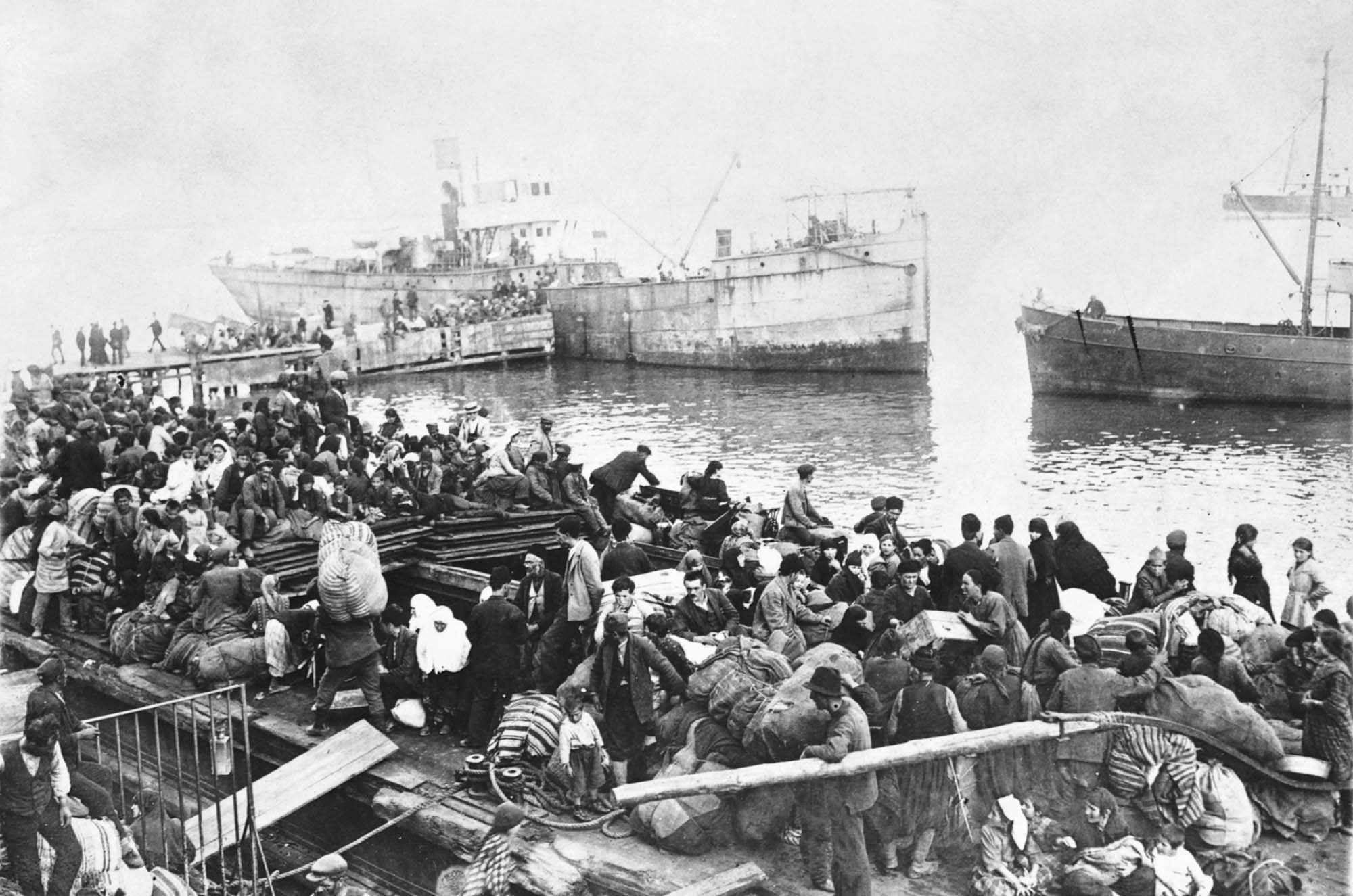 Black and white photo of refugees standing on a dock awaiting departure by three boats, in the background.